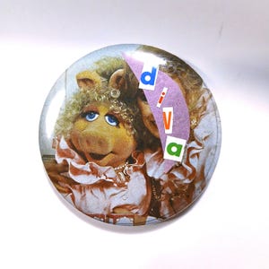 May include: Round button featuring Miss Piggy from The Muppets. The button has a light pink and white color scheme, with the word "DIVA" in colorful letters. Miss Piggy is wearing a pink dress and a pearl necklace.