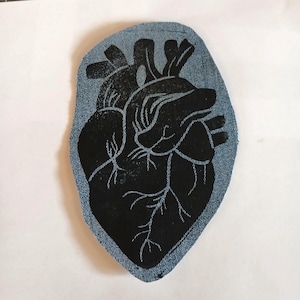 May include: A denim fabric patch featuring a black screen-printed anatomical heart design. The patch has a light blue denim background with a visible weave texture and a dark, solid black ink illustration of the heart with detailed veins.