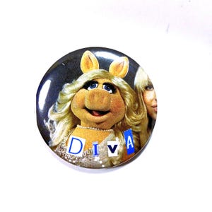 May include: A round button featuring Miss Piggy from The Muppets, with the word "DIVA" in block letters. Miss Piggy is depicted with blonde hair, blue eyes, and a pearl necklace. The button has a glossy finish and a white background.