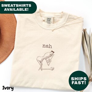 Könnte beinhalten: Elfenbeinfarbenes Sweatshirt mit Rundhalsausschnitt. Das Shirt zeigt eine Cartoon-Illustration einer Kreatur mit Sonnenbrille und dem Wort "nah" darüber. Auf dem Shirt stehen die Worte "Sweatshirts Available!" und "Ships Fast!"