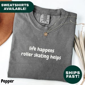 Puede incluir: Una camiseta gris con el texto blanco "life happens roller skating helps". La camiseta está doblada y tiene un collar encima. La camiseta está etiquetada como "Pepper" y "Comfort Colors".