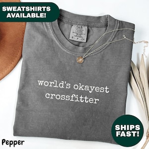 May include: A gray t-shirt with the text "world's okayest crossfitter" in white. The shirt is folded and has a crew neck. A gold necklace with a pendant rests on the shirt. The shirt is labeled "Pepper".