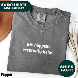May include: A gray Comfort Colors t-shirt with the text "life happens creativity helps" in white. A gold necklace with a pendant rests on the shirt. The shirt is folded and has a tag that says "S - P - CH".