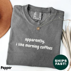 May include: A gray t-shirt with the text "apparently, i like morning coffees" in white. The shirt is folded and has a necklace draped over it. A brown hat and dried leaves are in the background.