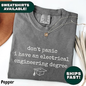 Puede incluir: Una camiseta gris con el texto "don't panic I have an electrical engineering degree" en blanco. Un gráfico de gorro de graduación está debajo del texto. Un collar dorado está sobre la camiseta. La camiseta está doblada sobre una superficie blanca.