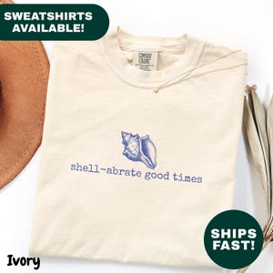 May include: Ivory-colored sweatshirt with a blue seashell graphic and the phrase "shell-abrate good times." The sweatshirt is a Comfort Colors brand and has a crew neck. The text "SWEATSHIRTS AVAILABLE!" is in a green box.