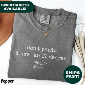 May include: A grey t-shirt with the text "don't panic I have an IT degree" and a graduation cap graphic. The shirt is folded and has a Comfort Colors tag. A gold necklace is draped across the shirt.
