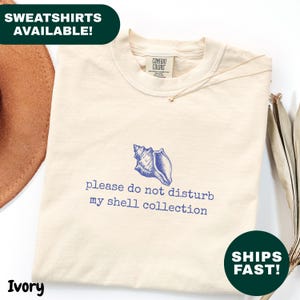 Può includere: Felpa color avorio con grafica a conchiglia blu e la scritta "please do not disturb my shell collection." Prodotta da Comfort Colors.