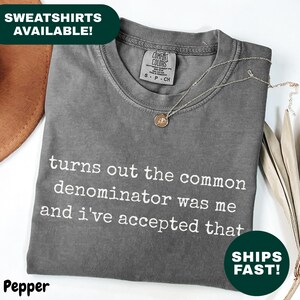 Könnte beinhalten: Ein graues T-Shirt mit dem weißen Text "turns out the common denominator was me and i've accepted that". Eine goldene Halskette mit Anhänger liegt auf dem Shirt. Das Shirt ist gefaltet und hat ein Comfort Colors-Etikett.