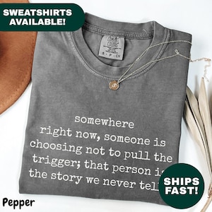 May include: A gray Comfort Colors t-shirt with the text "somewhere right now, someone is choosing not to pull the trigger; that person is the story we never tell." A gold necklace is draped over the shirt.