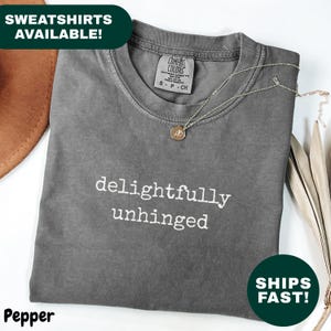 May include: A gray t-shirt with the words "delightfully unhinged" in white. The shirt is folded and has a necklace draped across it. The tag reads "Comfort Colors". The word "Pepper" is at the bottom left.