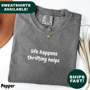 May include: A folded, pepper-colored t-shirt with the phrase "life happens thrifting helps" in white text. A gold-colored necklace rests on the shirt. The words "SWEATSHIRTS AVAILABLE!" and "SHIPS FAST!" are also visible.