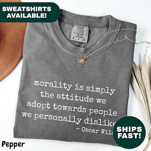 以下が含まれることがあります： 「道徳とは、私たちが個人的に嫌いな人々に対してとる態度にすぎない - オスカー・ワイルド」という引用句が白で書かれたグレーのTシャツ。 金色のネックレスがシャツの上にあります。