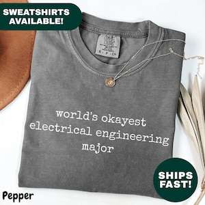 May include: A grey t-shirt with the text "world's okayest electrical engineering major" in white. A necklace rests on the folded shirt. The shirt is labelled "Pepper".