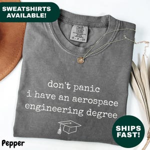 Pode incluir: Uma camiseta cinza com o texto "don't panic i have an aerospace engineering degree" em branco. Um gráfico de chapéu de formatura está abaixo do texto. Um colar de ouro está sobre a camiseta.
