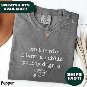 Może przedstawiać: Szara koszulka z białym napisem "don't panic I have a public policy degree". Poniżej tekstu znajduje się grafika czapki absolwenta. Złoty naszyjnik spoczywa na koszulce. Koszulka jest złożona i na białej powierzchni.
