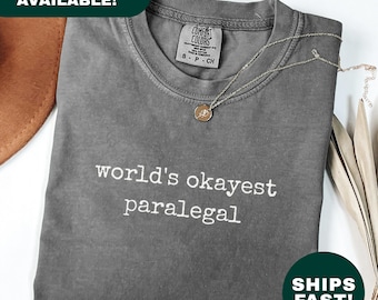 May include: A gray t-shirt with the text "world's okayest paralegal" in white. The shirt is folded and has a Comfort Colors tag. A gold necklace with a pendant rests on the shirt. The word "Pepper" is at the bottom left.