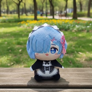 Könnte beinhalten: Plüschpuppe einer Figur mit blauen Haaren, blauen Augen und einer rosa Schleife. Die Puppe trägt ein schwarz-weißes Dienstmädchen-Outfit. Die Puppe sitzt auf einer Holzoberfläche mit einem verschwommenen Hintergrund eines Parks mit Bäumen und Blumen.