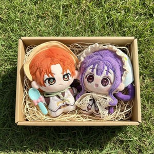 May include: Two plush dolls with large eyes and detailed embroidered features sit in a cardboard box filled with straw. One doll has orange hair and a hat, the other has purple hair and a bonnet. A blue spoon is included.