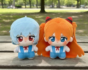 May include: Two plush dolls, one with light blue hair and the other with orange hair, both with detailed facial features and wearing white shirts and blue overalls. The dolls are sitting on a wooden surface with a blurred green background.