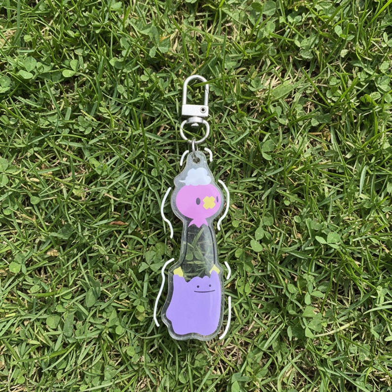 Pode incluir: Um porta-chaves de acr&iacute;lico transparente com um design de personagem de desenho animado em rosa e roxo. O porta-chaves tem um fecho prateado e um contorno branco. A personagem tem uma cabe&ccedil;a rosa, uma flor amarela e um corpo roxo. O porta-chaves est&aacute; na relva verde.