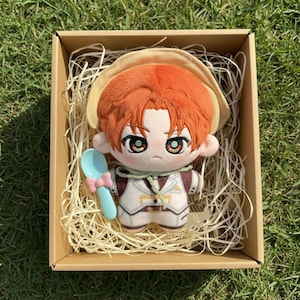 May include: A plush doll with orange hair and a tan hat, wearing a white vest with brown accents, sits in a cardboard box filled with straw. A light blue spoon with a pink bow is included. The doll has large eyes and is designed to look like a character.