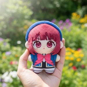 May include: A plush doll with a cartoon-like design. The doll has red hair, large pink eyes, and a blue headband. It wears a blue jacket with a pink bow and a black and white outfit. The doll is held in a hand against a blurred background of greenery.