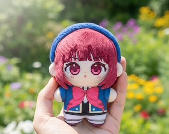 May include: A plush doll with a cartoon-like design. The doll has red hair, large pink eyes, and a blue headband. It wears a blue jacket with a pink bow and a black and white outfit. The doll is held in a hand against a blurred background of greenery.