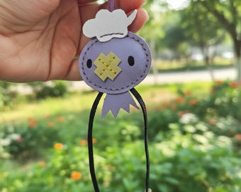 May include: A handmade purple leather keychain featuring a cartoon character design. The character has a white cloud-shaped hat, a yellow cross-shaped design, and black eyes. It has a black string attached for hanging.