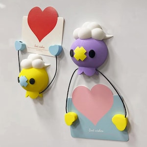 Op de afbeelding: Twee grillige wanddecoraties. De ene is geel, de andere paars, elk met een cartoongezicht en een kaart met een hart. Op de kaarten staat "Best wishes". De decoraties zijn met kleine blauwe hartjes aan de muur bevestigd.