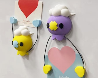 May include: Two whimsical wall decorations. One is yellow, the other purple, each with a cartoonish face and holding a card with a heart. The cards have the words "Best wishes" printed on them. The decorations are attached to the wall with small blue hearts.