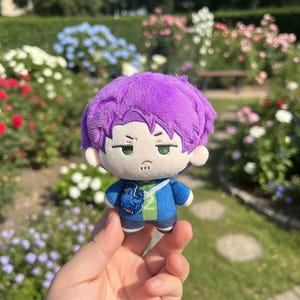 May include: A plush doll with purple hair, green eyes, and a blue jacket. The doll has a neutral expression and is held in front of a garden with colorful flowers. The doll's outfit includes a blue jacket and dark blue shorts.