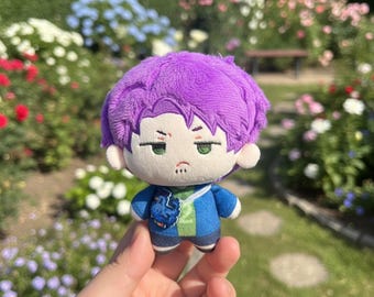 May include: A plush doll with purple hair, green eyes, and a blue jacket. The doll has a neutral expression and is held in front of a garden with colorful flowers. The doll's outfit includes a blue jacket and dark blue shorts.