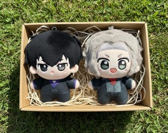 May include: Two plush dolls in a cardboard box. One doll has black hair and a black suit with a purple collar. The other doll has gray hair and a gray suit with a red collar. The dolls have large eyes and are sitting on straw.
