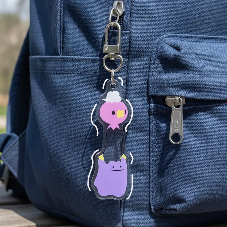 Pode incluir: Um chaveiro de acr&iacute;lico transparente com dois personagens de desenho animado. O personagem superior &eacute; rosa com uma nuvem branca e detalhes amarelos. O personagem inferior &eacute; roxo com um rosto sorridente. O chaveiro &eacute; preso a um fecho prateado.