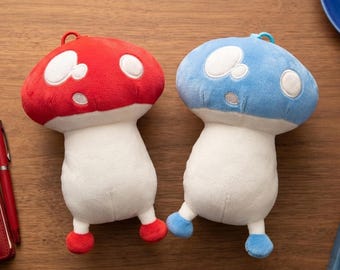 May include: Two plush mushroom toys, one with a red cap and one with a blue cap. Both have white stems, white spots on their caps, and small rounded feet matching their cap colors. Each mushroom features a small loop at the top for hanging.