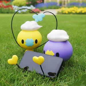 Puede incluir: Dos figuras esféricas de dibujos animados, una amarilla y otra morada, con sombreros blancos en forma de nube y ojos negros, están sobre hierba verde. Un smartphone negro está apoyado con soportes amarillos en forma de corazón. Un alambre negro forma un corazón sobre la figura amarilla.