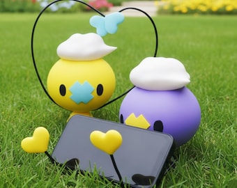 May include: Two spherical, cartoon-like figures, one yellow and one purple, with white cloud-shaped hats and black eyes, stand on green grass. A black smartphone is propped up with yellow heart-shaped stands. A black wire forms a heart shape above the yellow figure.