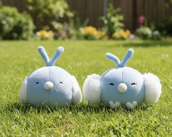 May include: Two light blue plush bird toys with white accents and small beaks sit on green grass. Each bird has a V-shaped head crest and fluffy white ear-like features. The toys are in a sunny outdoor setting.