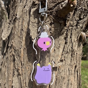 Pode incluir: Um chaveiro de acr&iacute;lico transparente com dois personagens de desenho animado. O personagem superior &eacute; rosa com uma nuvem branca e uma cruz amarela. O personagem inferior &eacute; roxo com um rosto sorridente e m&atilde;os amarelas. O chaveiro tem um fecho prateado.