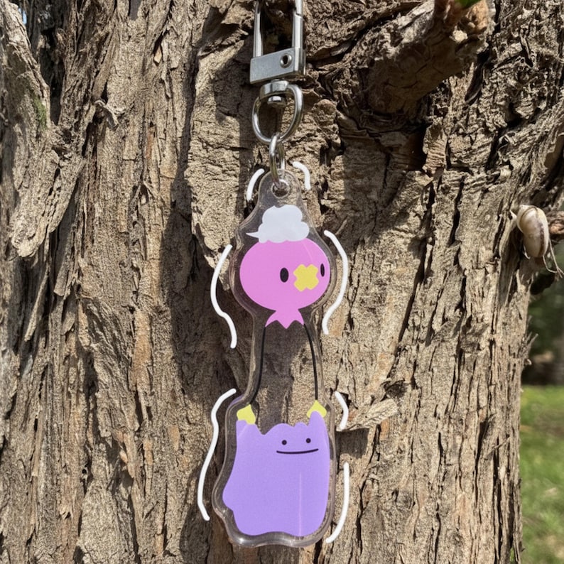 Pode incluir: Um chaveiro de acr&iacute;lico transparente com dois personagens de desenho animado. O personagem superior &eacute; rosa com uma nuvem branca e uma cruz amarela. O personagem inferior &eacute; roxo com um rosto sorridente e m&atilde;os amarelas. O chaveiro tem um fecho prateado.