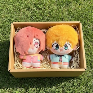 May include: Two plush dolls with large eyes and colorful outfits are nestled in a cardboard box filled with straw. One doll has pink hair and a pink outfit, the other has yellow hair and a blue outfit.