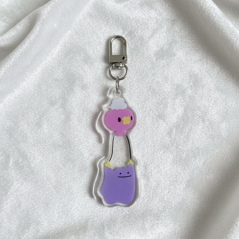 Pode incluir: Um chaveiro de acr&iacute;lico transparente com um personagem Ditto roxo segurando um bal&atilde;o rosa com uma nuvem branca. O chaveiro tem um fecho prateado para f&aacute;cil fixa&ccedil;&atilde;o. Um acess&oacute;rio fofo e divertido.