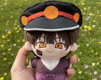 May include: A plush doll featuring brown hair and orange eyes, wearing a black military-style cap with an orange band and circular emblem. The doll has a small black square patch with red Japanese characters on its right cheek and wears a purple outfit.