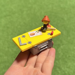 Könnte beinhalten: Ein Miniatur-gelber Werkbank mit silberner Werkzeugkastenbasis. Die Werkbank hat eine kleine Lampe, einen Hammer, einen Schraubendreher und eine Zeichnung. Ein silbernes L-förmiges Lineal befindet sich an der Seite.