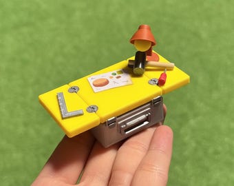 May include: A miniature yellow workbench with a silver toolbox base. The workbench has a small lamp, a hammer, a screwdriver, and a drawing. A silver L-shaped ruler is on the side.