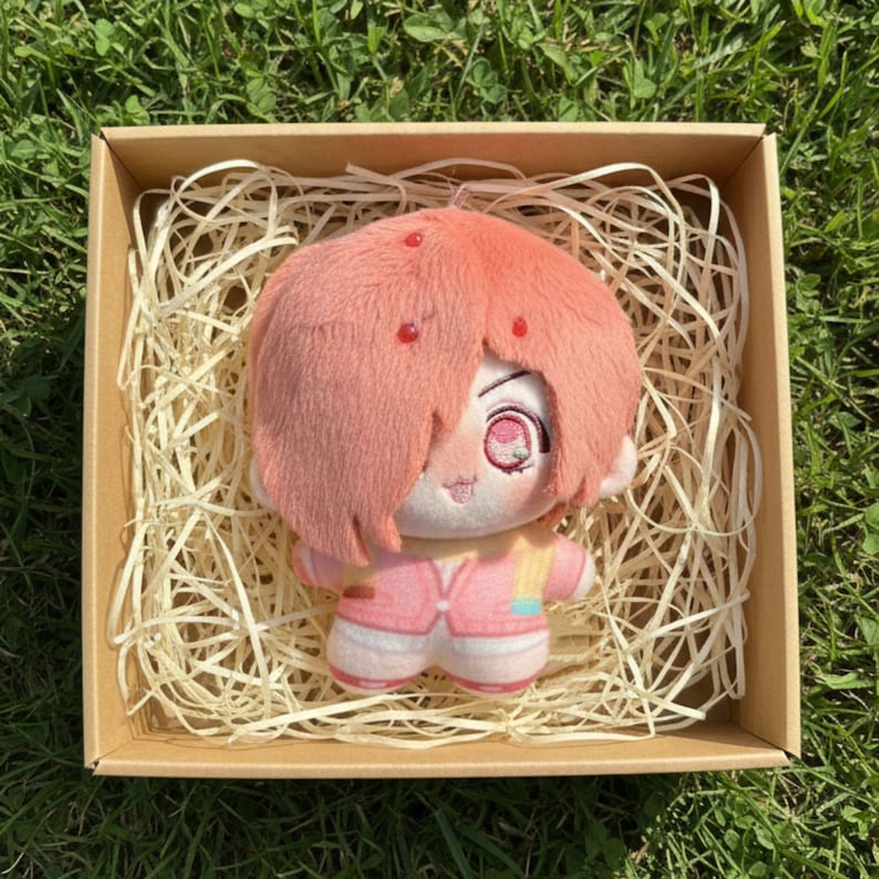 May include: A plush doll with pink hair and a pink outfit, presented in a brown cardboard box filled with straw. The doll has a detailed face with large eyes and is designed in a cartoon style. The doll is a collectible item.