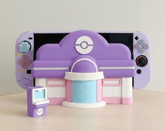 May include: A purple and white Nintendo Switch case with a building design, a small arcade machine, and matching controller buttons. The building has a purple roof with a white logo and a blue and white door. The controller has a black joystick.