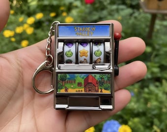 May include: Miniature silver-colored slot machine keychain featuring the text "Stardew Valley." The reels display various game icons. The bottom panel shows a landscape scene with a red barn. A red handle is on the side.
