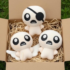 May include: Three plush figures with large black eyes and small arms are nestled in a cardboard box filled with straw. One figure has a black eye patch. The figures are a light beige color.
